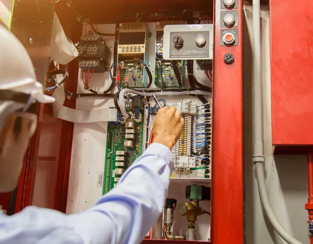 A technician wearing a hard hat and safety glasses is inspecting or repairing a fire alarm control panel. The panel contains various electronic components, wiring, and circuit boards. The person is using a tool to check the internal systems, indicating routine maintenance or troubleshooting of the fire protection system.