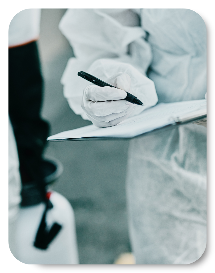 Close-up of an inspector in full protective gear conducting an asbestos survey, writing notes on a clipboard while holding a pen. A spray canister is partially visible in the background.