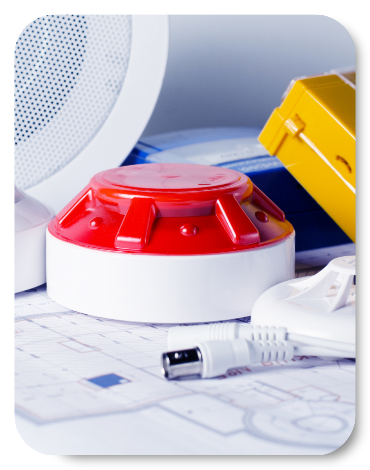 Close-up of fire alarm components, including a red fire detector, wiring, and safety equipment, placed on top of architectural floor plans during a Fire Risk Assessment.