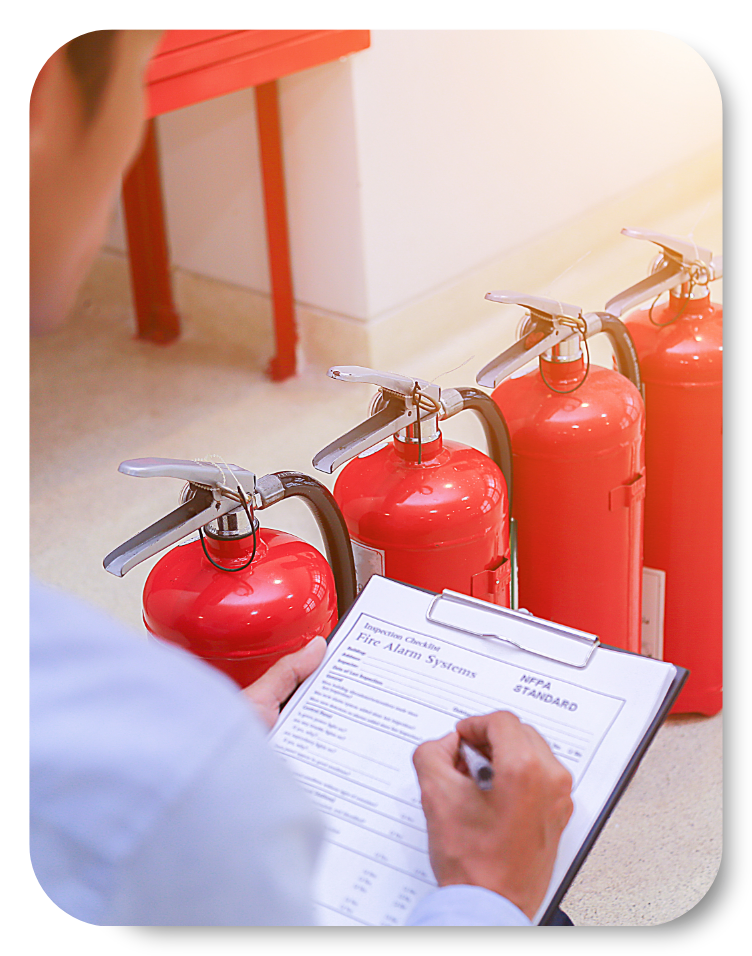 Inspector conducting a Fire Risk Assessment, reviewing and completing a fire alarm systems checklist on a clipboard in front of a row of red fire extinguishers.