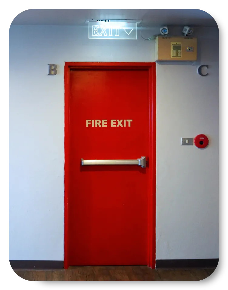 Gray emergency exit door with a panic bar, red fire alarm, and fire sprinkler piping on a concrete wall.