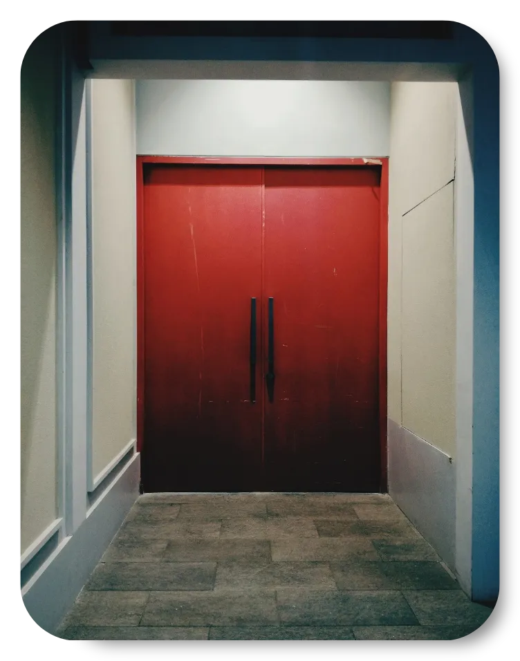 Double red metal doors in a dimly lit hallway with tiled flooring and beige walls.
