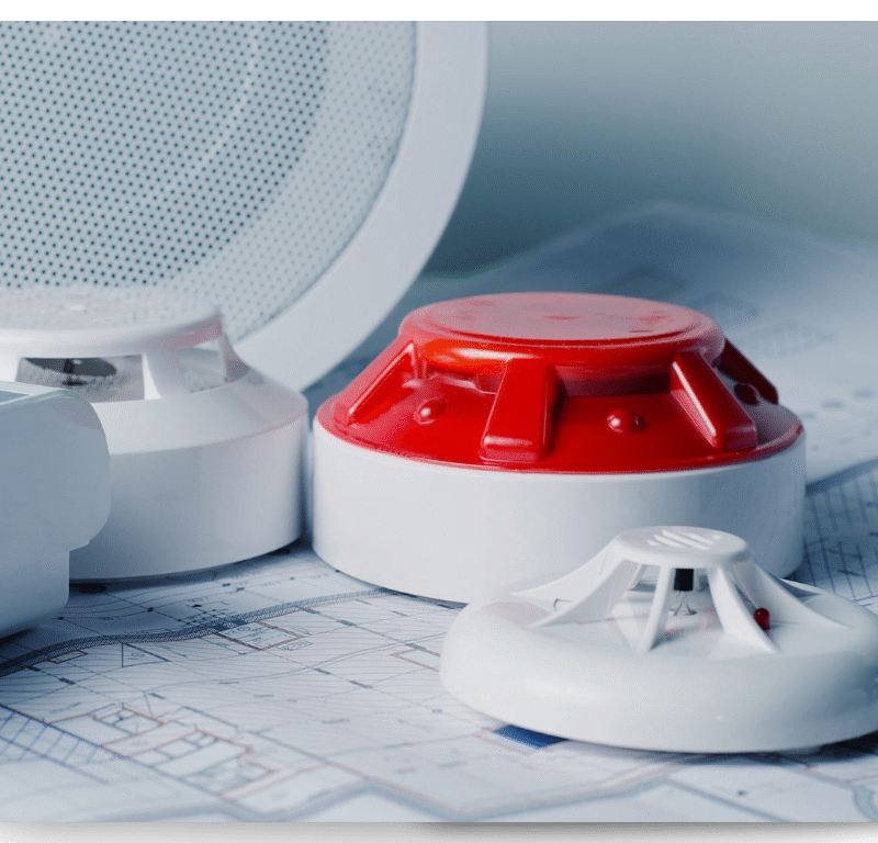 Fire alarm devices including a smoke detector, heat detector, and alarm sounder placed on top of architectural blueprints.