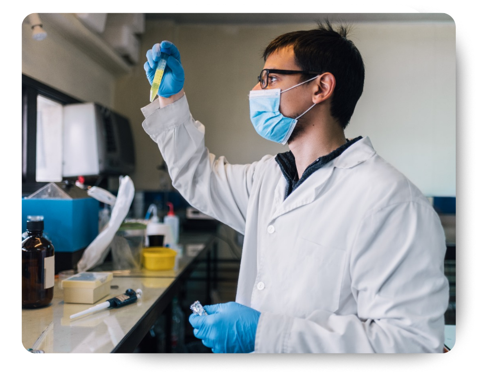 A male scientist wearing a lab coat, gloves, and a face mask inspects a test tube filled with liquid in a laboratory.