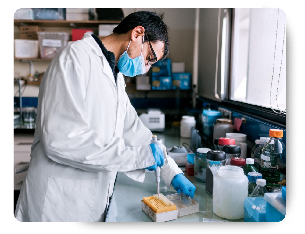 A scientist wearing protective eyewear, a face mask, and gloves carefully pouring liquid from a pipette into a container in a laboratory.