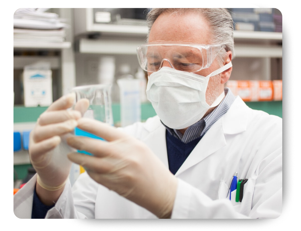 A scientist wearing safety goggles, a face mask, and gloves examines a beaker containing blue liquid in a laboratory.